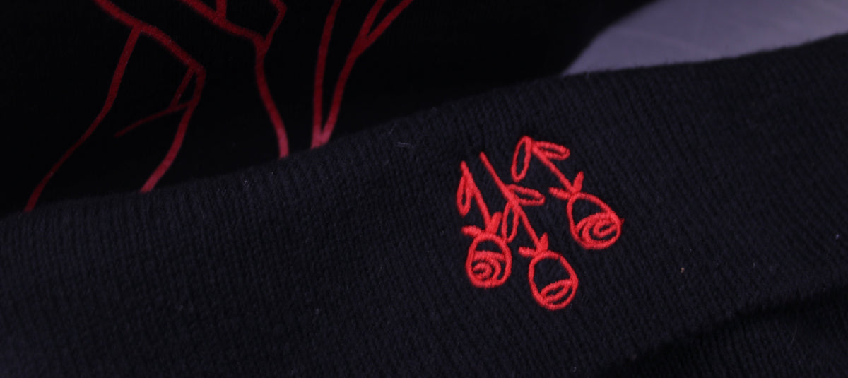 Close-up of a black knit beanie hat featuring red embroidered details, including a small floral embroidered patch on the cuff and a larger abstract line design on a separate piece of apparel.